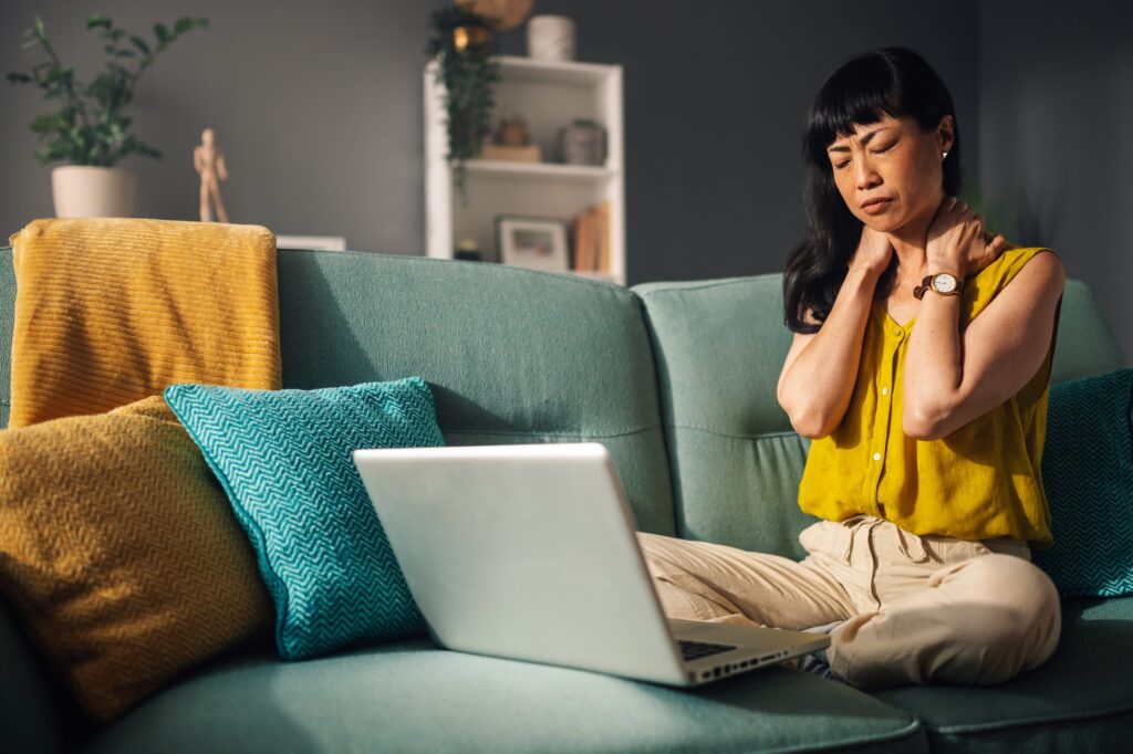 Asian woman experiencing neck pain while using laptop on her couch
